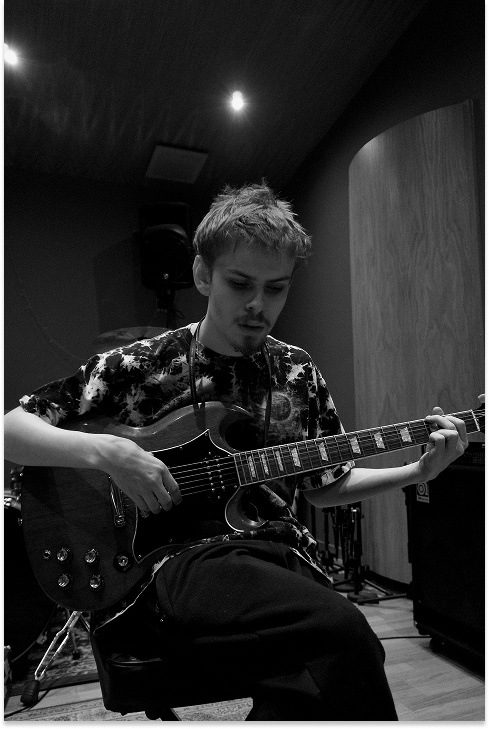 A black-and-white photograph of a young man (Marshall) playing an electric guitar in a professional recording studio, seated with a focused expression, surrounded by amplifiers and studio equipment.