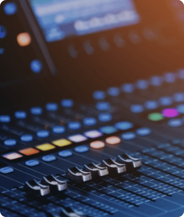 A close-up of a professional mixing console with illuminated faders, colourful buttons, and control knobs, with a warm ambient lighting effect.
